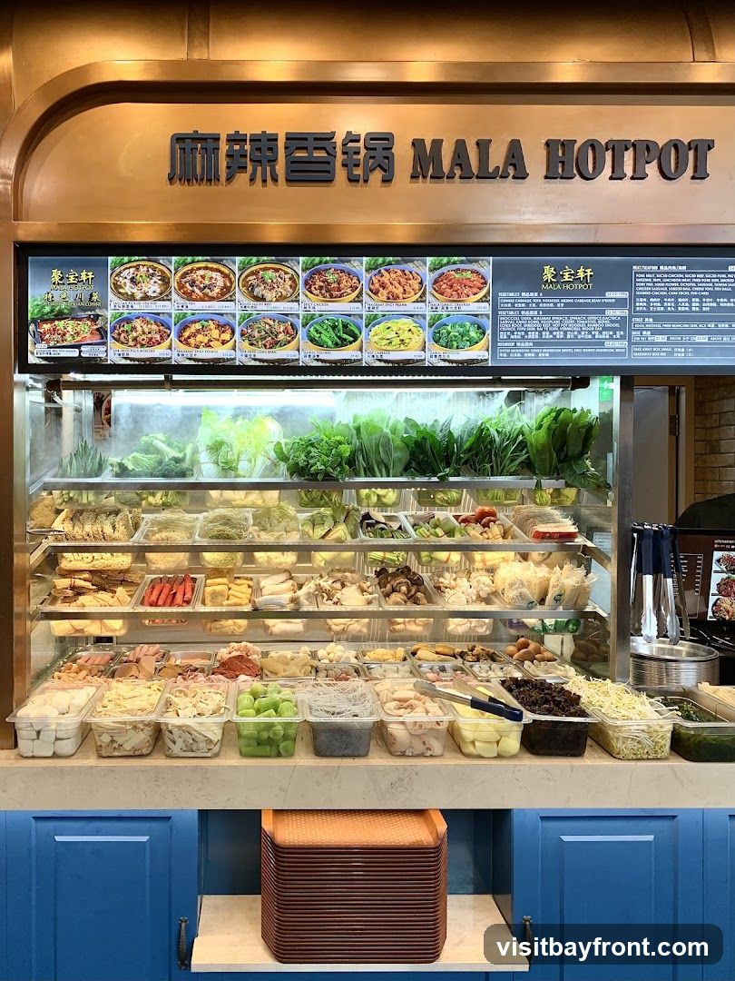 A display case filled with various ingredients for Mala Hotpot, including fresh vegetables, meats, tofu, and noodles. Above the display is a sign with the words "MALA HOTPOT" and a menu board showing pictures of dishes and prices.