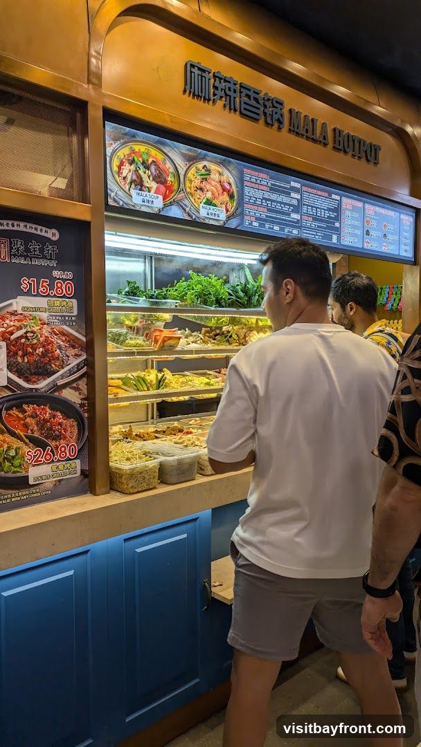 A display case filled with various ingredients for mala hotpot, including fresh vegetables, meats, and seafood. A menu board is visible above the display, featuring images of dishes and prices. Several customers are standing in front of the display, looking at the options.