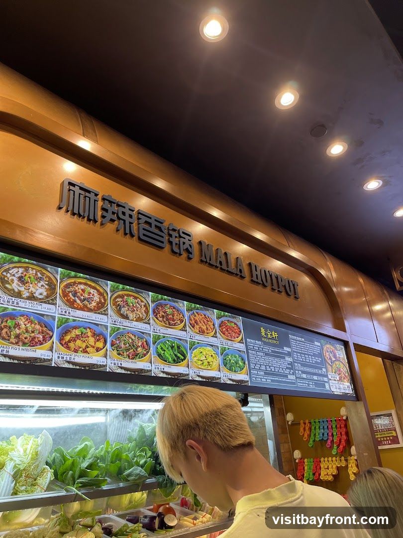 A person is looking at a display of fresh vegetables and a menu board for Mala Hotpot. The menu board features images of various dishes and their names, along with prices. The restaurant's name, "MALA HOTPOT," is visible in large lettering above the menu.