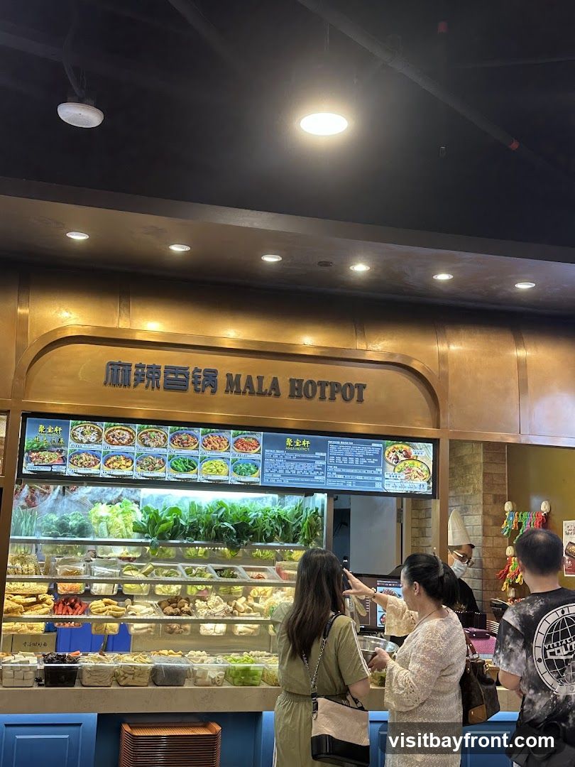 A display of fresh ingredients for mala hotpot, including various vegetables, meats, and tofu products. Customers are seen selecting their ingredients. A menu board above shows pictures of dishes and prices.