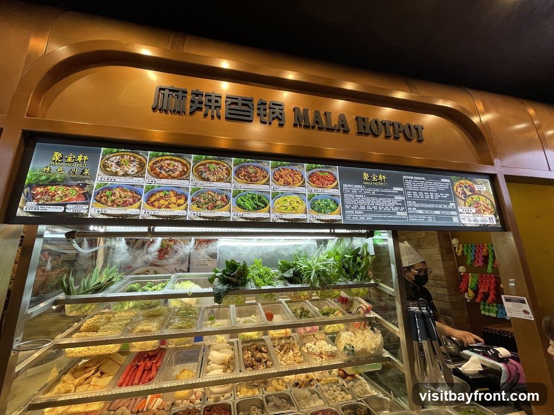 A display case filled with various ingredients for mala hotpot, including vegetables, meats, and noodles. Above the display is a menu board with pictures of dishes and text in Chinese and English, featuring the name "MALA HOTPOT". A staff member wearing a hat and mask is visible behind the counter.