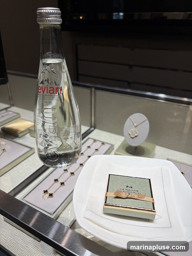 A display case featuring several necklaces with clover-shaped pendants. A bottle of Evian water stands upright in the center, and a small box of chocolates from 'La Maison du Chocolat' is presented on a white napkin in a white dish. The overall scene suggests a luxury retail environment.