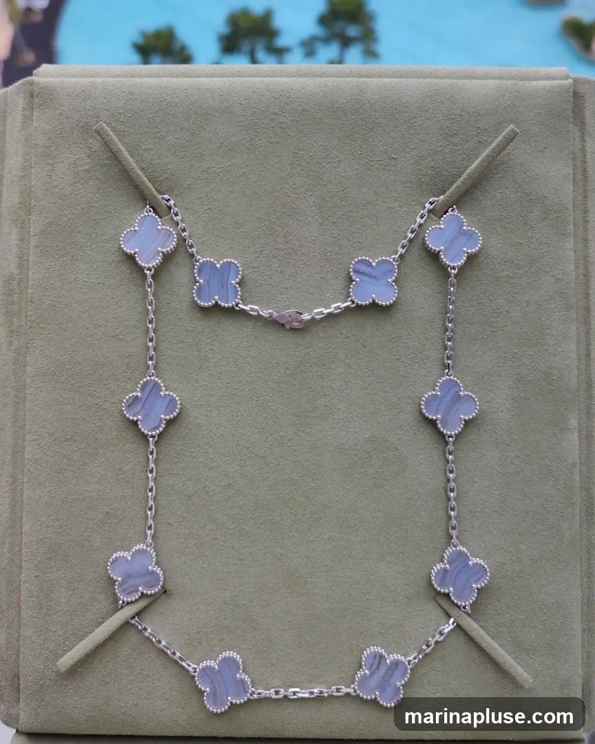 A silver necklace with multiple clover-shaped charms featuring a blue and white striped stone. The necklace is displayed on a green velvet stand. In the background, out of focus, is a body of water and trees.