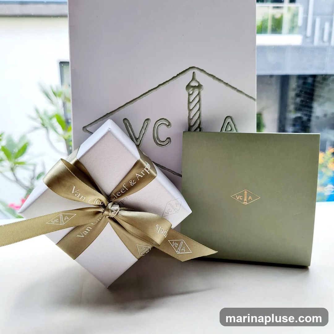 A white gift box with a gold ribbon tied around it sits in front of a larger white box with the Van Cleef & Arpels logo embossed on it. To the right is a small green box with the Van Cleef & Arpels logo.
