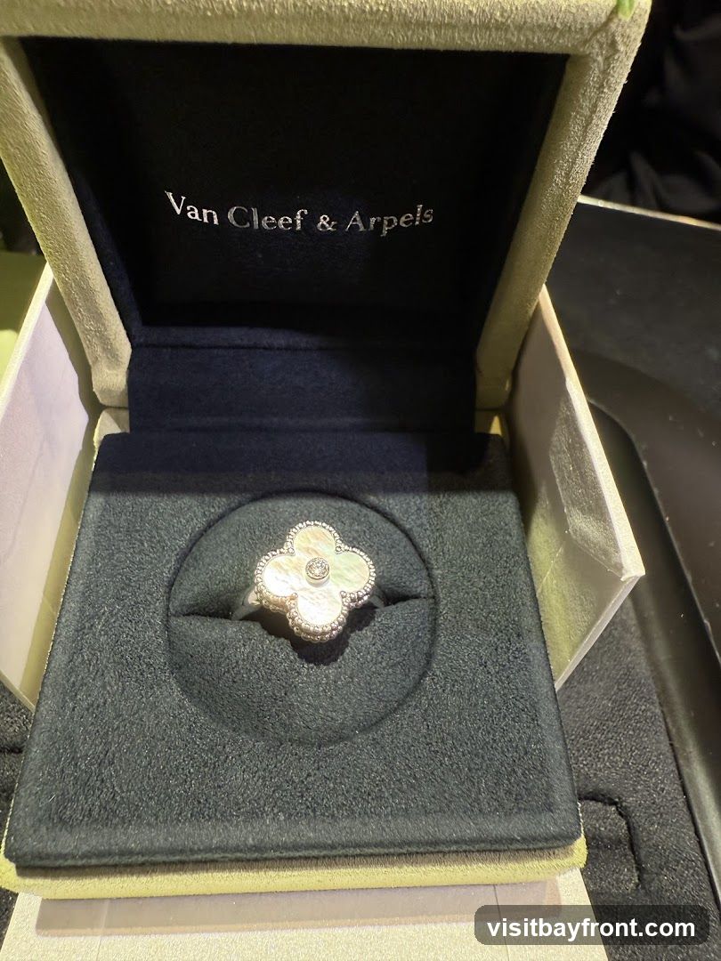 A Van Cleef & Arpels ring with a mother-of-pearl clover design and a diamond center is displayed in its velvet-lined box. The inside lid of the box features the brand's logo, "Van Cleef & Arpels," in silver lettering.