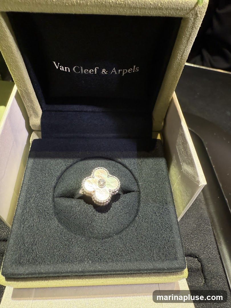 A Van Cleef & Arpels ring with a mother-of-pearl clover design and a diamond center is displayed in its velvet-lined box. The inside lid of the box features the brand's logo, "Van Cleef & Arpels," in silver lettering.