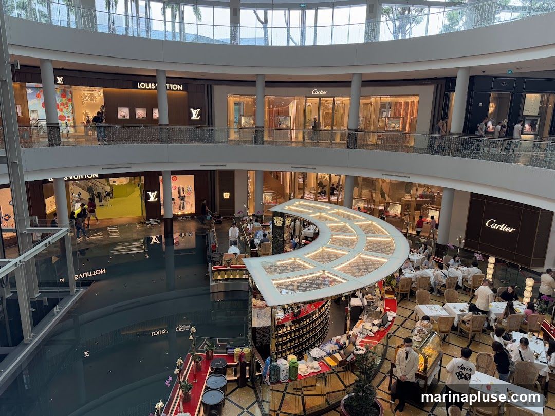 An aerial view of a multi-level indoor shopping mall. On the upper levels, storefronts for luxury brands like Louis Vuitton and Cartier are visible, with displays of merchandise. Below, a circular cafe or restaurant area is set up with tables and chairs, where patrons are seated. A body of water, possibly a canal or reflecting pool, is present in the lower section of the mall.