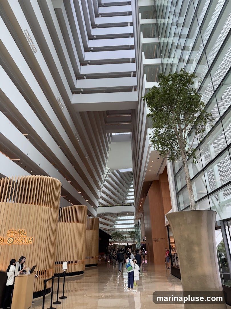 A wide shot of a modern hotel atrium with a tree in the center. The atrium has multiple levels of balconies overlooking a central walkway. On the left, there is a reception area with wooden paneling and a sign that reads "BLOSSOM". People are walking in the atrium.