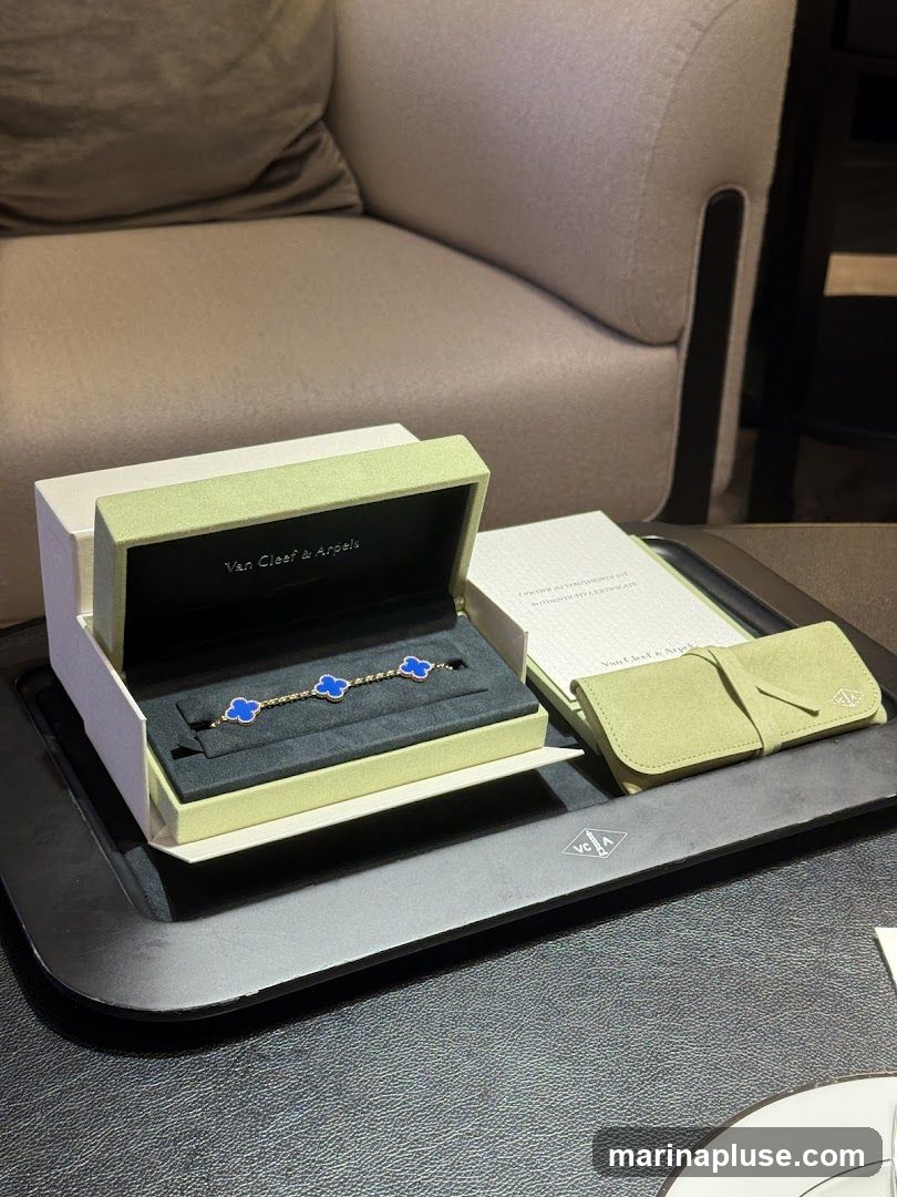 A Van Cleef & Arpels bracelet with blue clover motifs is displayed in its opened box. Next to it is a green pouch and a certificate of authenticity. The items are placed on a black tray, with a plush chair in the background.