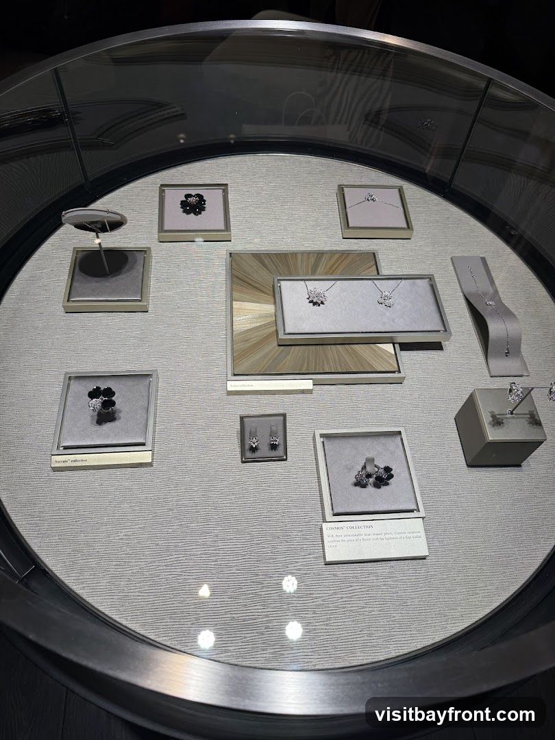 A display case filled with various pieces of jewelry, including necklaces, earrings, and bracelets. The jewelry is arranged on grey velvet stands and in framed displays. Some pieces are floral-themed, while others are more geometric. A small plaque reads "COSMOS COLLECTION" and describes the pieces as "delicate, more vulnerable heart-shaped petals. Cosmos creations capture the grace of a flower with the lightness of a fine-loaded feather."