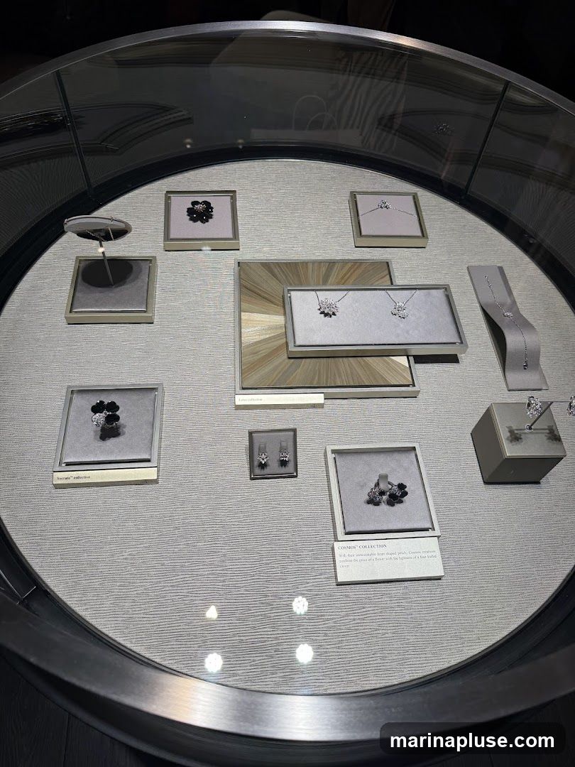 A display case filled with various pieces of jewelry, including necklaces, earrings, and bracelets. The jewelry is arranged on grey velvet stands and in framed displays. Some pieces are floral-themed, while others are more geometric. A small plaque reads "COSMOS COLLECTION" and describes the pieces as "delicate, more vulnerable heart-shaped petals. Cosmos creations capture the grace of a flower with the lightness of a fine-loaded feather."