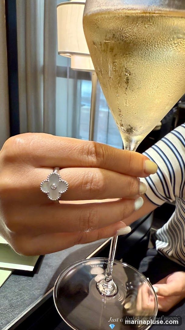 A close-up shot shows a woman's hand adorned with a white clover-shaped ring with a diamond center, likely from Van Cleef & Arpels. She is holding a champagne flute filled with a bubbly, golden beverage. The background is softly blurred, revealing a lamp with a cream-colored shade and draped curtains. The overall scene suggests a celebratory or luxurious setting.
