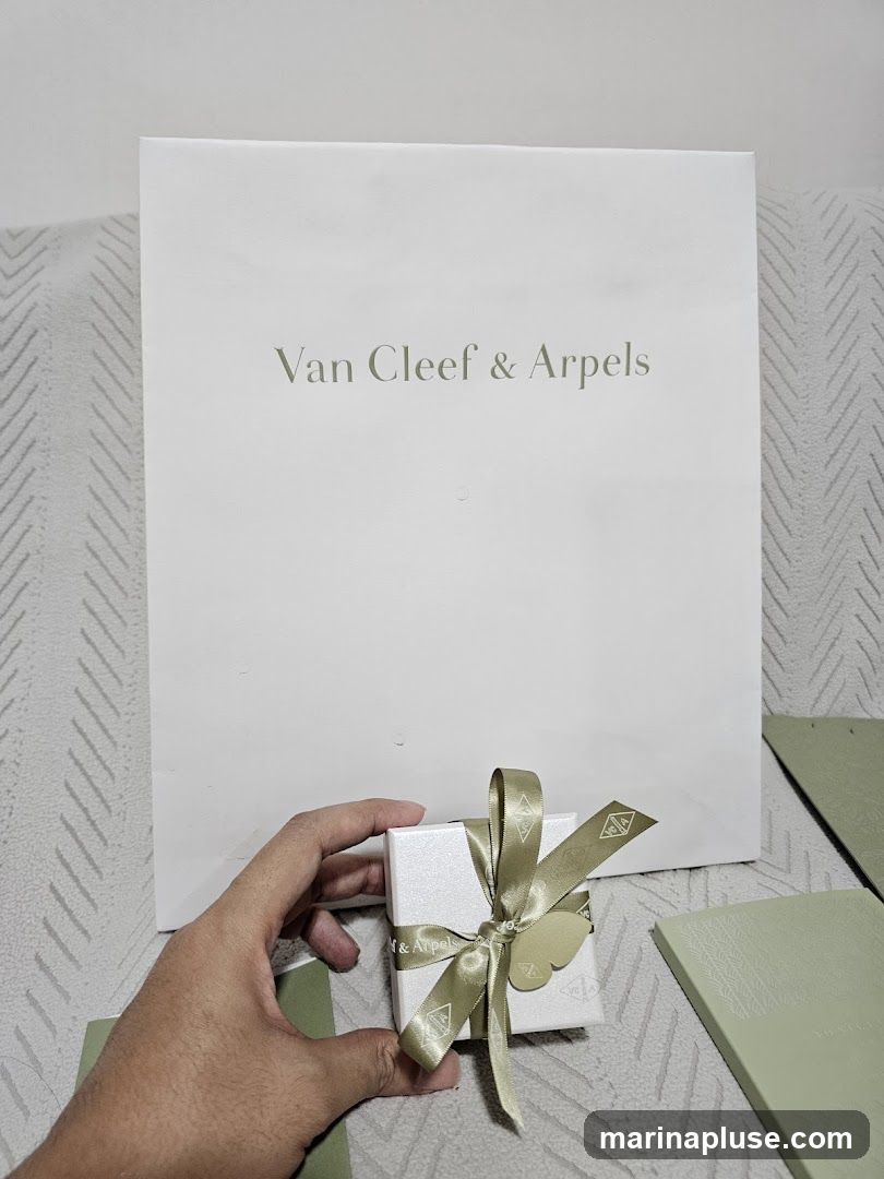 A hand holds a small, white, glittery gift box tied with a gold ribbon. The ribbon has the Van Cleef & Arpels logo printed on it. Behind the box is a large, white shopping bag with the words "Van Cleef & Arpels" printed in a light green font. To the right of the box are two light green booklets or folders, also with embossed designs. The items are placed on a textured surface, possibly a couch or blanket.