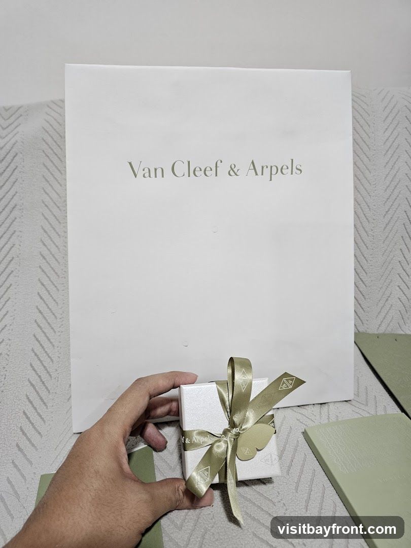 A hand holds a small, white, glittery gift box tied with a gold ribbon. The ribbon has the Van Cleef & Arpels logo printed on it. Behind the box is a large, white shopping bag with the words "Van Cleef & Arpels" printed in a light green font. To the right of the box are two light green booklets or folders, also with embossed designs. The items are placed on a textured surface, possibly a couch or blanket.