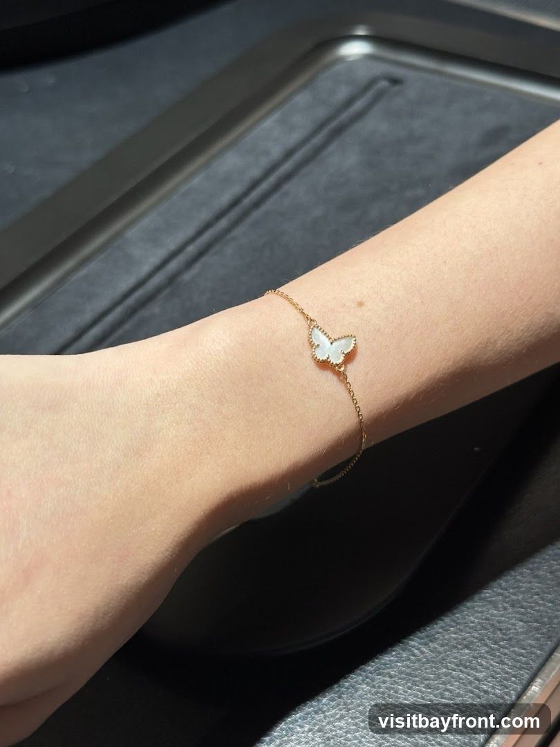 A close-up shot of a person's arm wearing a delicate gold bracelet with a white butterfly charm. The charm appears to be made of mother-of-pearl. The background is dark and textured, possibly a jewelry display case.