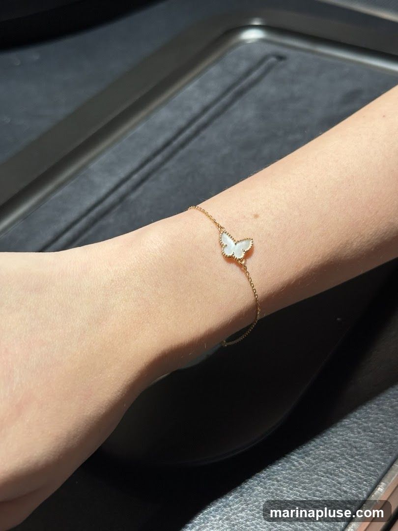 A close-up shot of a person's arm wearing a delicate gold bracelet with a white butterfly charm. The charm appears to be made of mother-of-pearl. The background is dark and textured, possibly a jewelry display case.