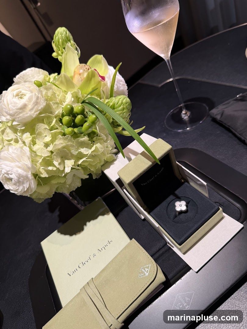 A white flower-shaped ring from Van Cleef & Arpels is displayed in its box, next to a bouquet of white and green flowers and a glass of champagne. A branded booklet and a pouch are also visible.