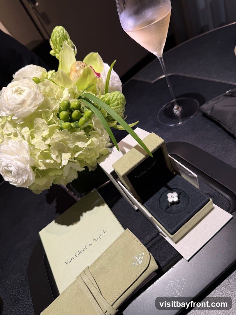 A white flower-shaped ring from Van Cleef & Arpels is displayed in its box, next to a bouquet of white and green flowers and a glass of champagne. A branded booklet and a pouch are also visible.