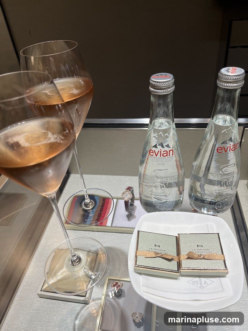 Two glasses of rose champagne are displayed next to two bottles of Evian water. In front of the water bottles are two small boxes of chocolates from La Maison du Chocolat, presented on a white dish. Several pieces of jewelry, including rings and a pendant, are visible in display cases below.
