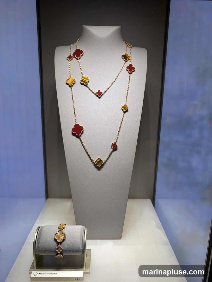 A display case featuring a long gold necklace and a bracelet, both adorned with clover-shaped motifs in red and tiger's eye stones. The necklace is draped over a grey mannequin bust, while the bracelet is presented on a grey stand. A small plaque below the bracelet reads "Alhambra® collection" and displays a logo.