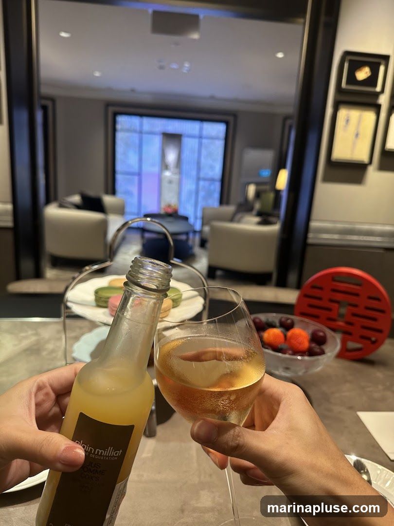 Two hands clink a bottle of juice and a glass of wine in a toast. In the background, a tiered stand holds colorful macarons, and a bowl of berries sits on the table. The setting appears to be an elegant lounge or dining area with comfortable seating and artwork on the walls.