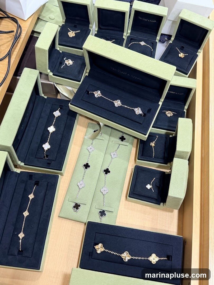 A high-angle shot showcases an assortment of Van Cleef & Arpels jewelry, primarily bracelets and necklaces, displayed in their signature light green boxes. The pieces feature the iconic four-leaf clover motif, with some made of gold and others adorned with white diamonds and mother-of-pearl or onyx inlays. The jewelry is arranged in an organized manner within open boxes and on display stands, suggesting a retail or display setting.