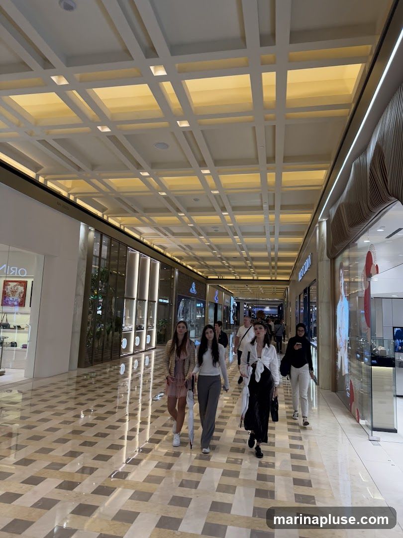 A group of young women and men are walking down a brightly lit corridor in a shopping mall. The ceiling features a geometric design with recessed lighting. The floor is tiled with a checkered pattern of light and dark marble. Storefronts with glass windows line both sides of the corridor, displaying merchandise.