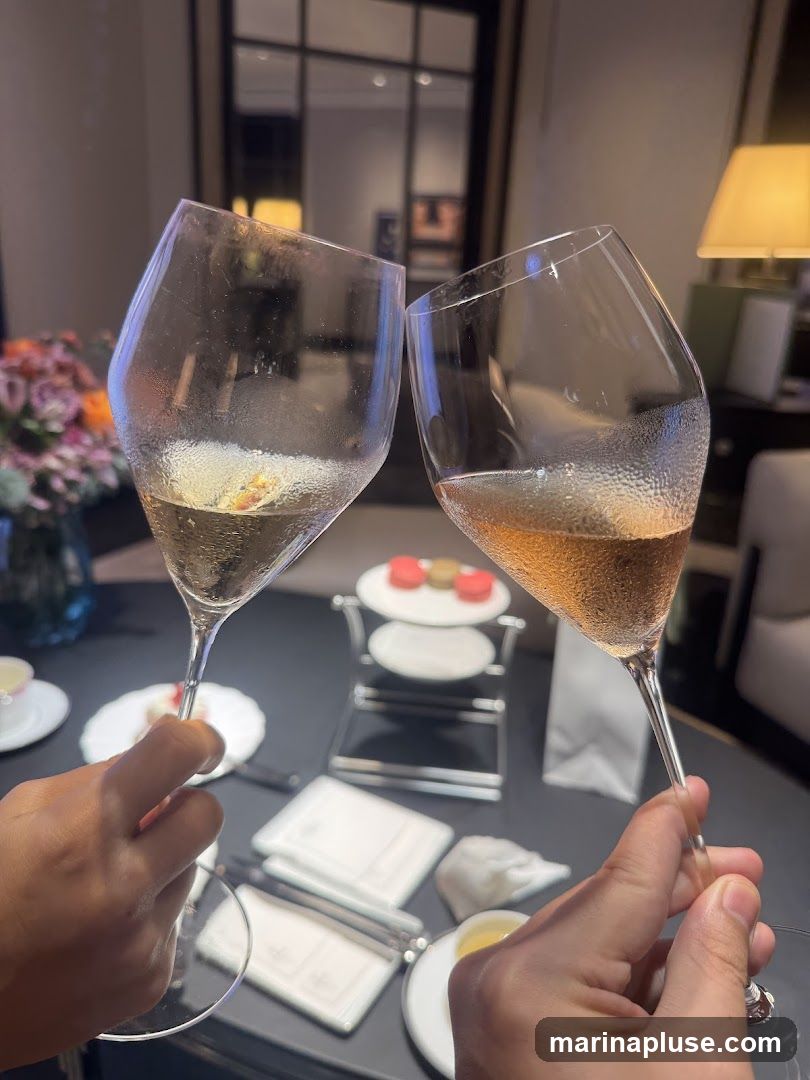 Two hands clink glasses of champagne in a toast. In the background, a tiered stand holds macarons, and a floral arrangement is visible. The setting appears to be an indoor lounge or dining area with a lamp and elegant decor.