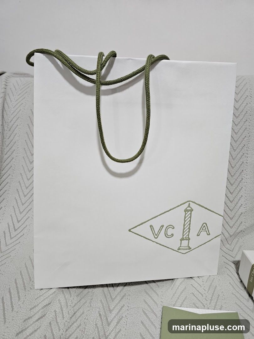 A white paper shopping bag with green rope handles and a green diamond-shaped logo containing the letters VCA and a candle illustration. A small white box and a green card are visible to the right of the bag.