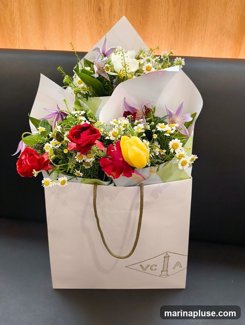 A light pink paper bag with a gold logo and rope handles contains a vibrant flower arrangement. The flowers include red roses, yellow tulips, white lilies, and small white daisies, with green foliage. The logo on the bag appears to be VC & A within a diamond shape, featuring a stylized lighthouse or column. The background is a dark surface with a wooden texture above.