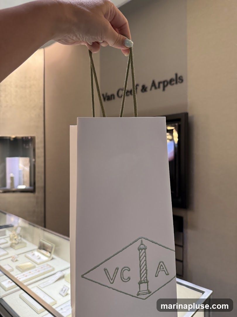A hand holds a white shopping bag with green handles. The bag has a green embossed logo featuring the letters VCA within a diamond shape, with a stylized lighthouse or column in the center. In the background, the words "Van Cleef & Arpels" are visible on the wall, along with display cases filled with jewelry.