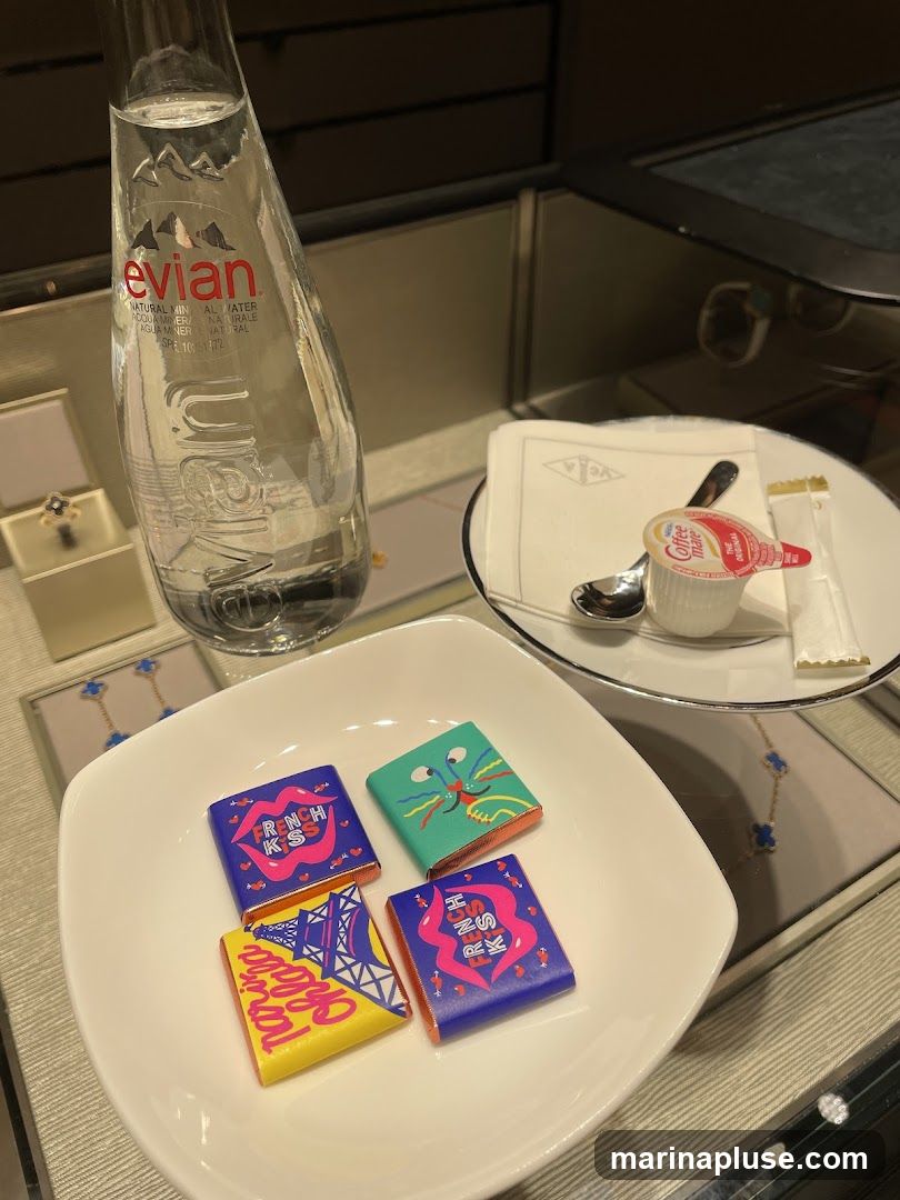 A white bowl holds four individually wrapped chocolates with colorful designs. One chocolate wrapper features a stylized Eiffel Tower and the text "Parisa". Another has a green background with a cartoon monster face. Two other chocolates have pink lips and the text "French Kiss" on a blue background. Next to the bowl is a bottle of Evian natural mineral water. A small white plate holds a packet of Coffee Mate creamer, a spoon, and a sugar packet, all on a white napkin with a logo. In the background, jewelry is displayed in cases.