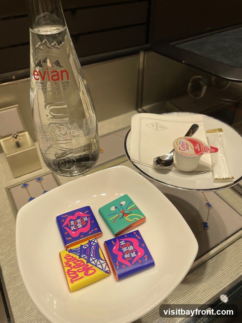 A white bowl holds four individually wrapped chocolates with colorful designs. One chocolate wrapper features a stylized Eiffel Tower and the text "Parisa". Another has a green background with a cartoon monster face. Two other chocolates have pink lips and the text "French Kiss" on a blue background. Next to the bowl is a bottle of Evian natural mineral water. A small white plate holds a packet of Coffee Mate creamer, a spoon, and a sugar packet, all on a white napkin with a logo. In the background, jewelry is displayed in cases.