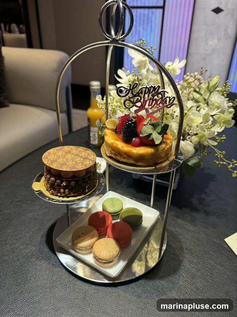 A tiered stand holds a selection of pastries and macarons, with a "Happy Birthday" topper on one of the cakes. A bottle of orange juice and a bouquet of white flowers are also visible.