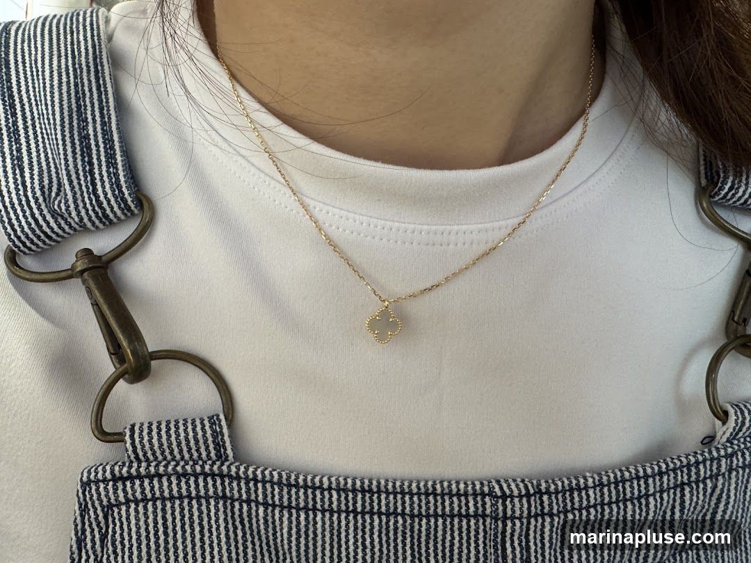 A close-up shot of a person wearing a delicate gold necklace with a small, white clover-shaped pendant. The person is also wearing a white t-shirt and blue and white striped overalls. The necklace is centered on the chest, and the pendant is clearly visible. The image focuses on the jewelry and the upper body of the person.