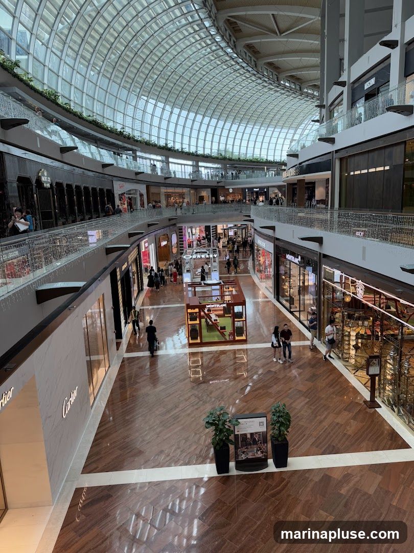A wide-angle view of the interior of a multi-level shopping mall with a distinctive glass roof. The mall features polished wooden floors, escalators, and various retail stores on different levels. People are seen browsing and walking through the mall. Prominent store names like Cartier and Montblanc are visible. There are also decorative displays and planters throughout the space.