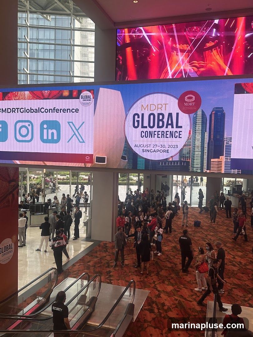 A large digital screen displays information about the MDRT Global Conference in Singapore, scheduled for August 27-30, 2023. The screen also shows social media icons for Facebook, Instagram, LinkedIn, and X. Below the screen, a crowd of people are gathered in a large indoor space with escalators and patterned carpeting.