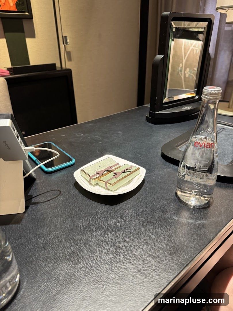 A close-up view of a dark, textured surface, likely a desk or table in a hotel room. On the surface, there are two small, elegantly wrapped gift boxes with ribbons, placed on a white square plate. To the right, a clear glass bottle of Evian water stands upright. In the background, a black vanity mirror is visible, reflecting a patterned wallpaper. A charging dock with a phone is also present on the left side of the frame.