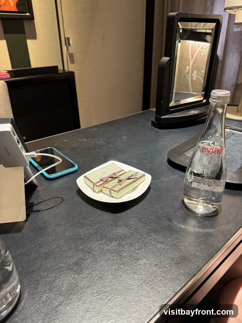 A close-up view of a dark, textured surface, likely a desk or table in a hotel room. On the surface, there are two small, elegantly wrapped gift boxes with ribbons, placed on a white square plate. To the right, a clear glass bottle of Evian water stands upright. In the background, a black vanity mirror is visible, reflecting a patterned wallpaper. A charging dock with a phone is also present on the left side of the frame.