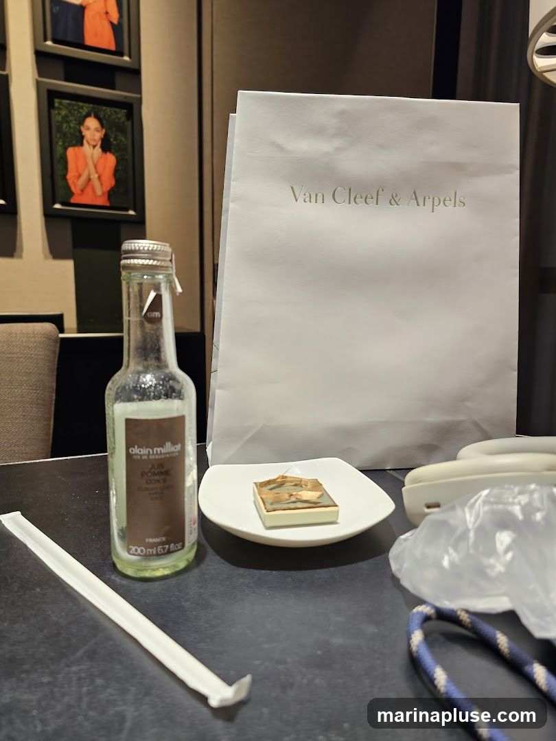 A white shopping bag with the Van Cleef & Arpels logo is placed on a table. Next to it is a bottle of Alain Milliat Jus Pomme Coq's drink and a small dessert on a white plate. A straw and a phone are also visible on the table. Framed pictures of a woman in an orange outfit are on the wall in the background.