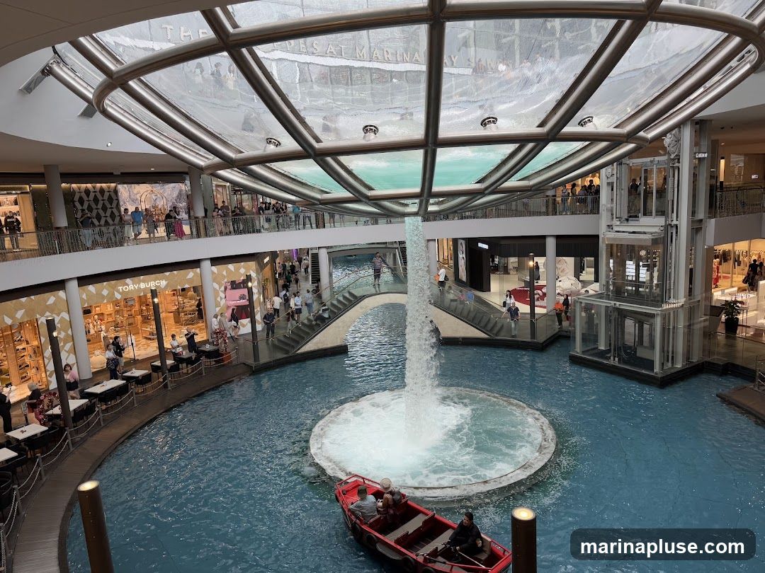 An interior view of a large, multi-level shopping mall. In the center is a circular pool of blue water with a fountain cascading from above. A small red boat with people in it is on the water. Surrounding the pool are shops and walkways. The ceiling is a large glass structure. People are seen walking around and browsing in the shops.