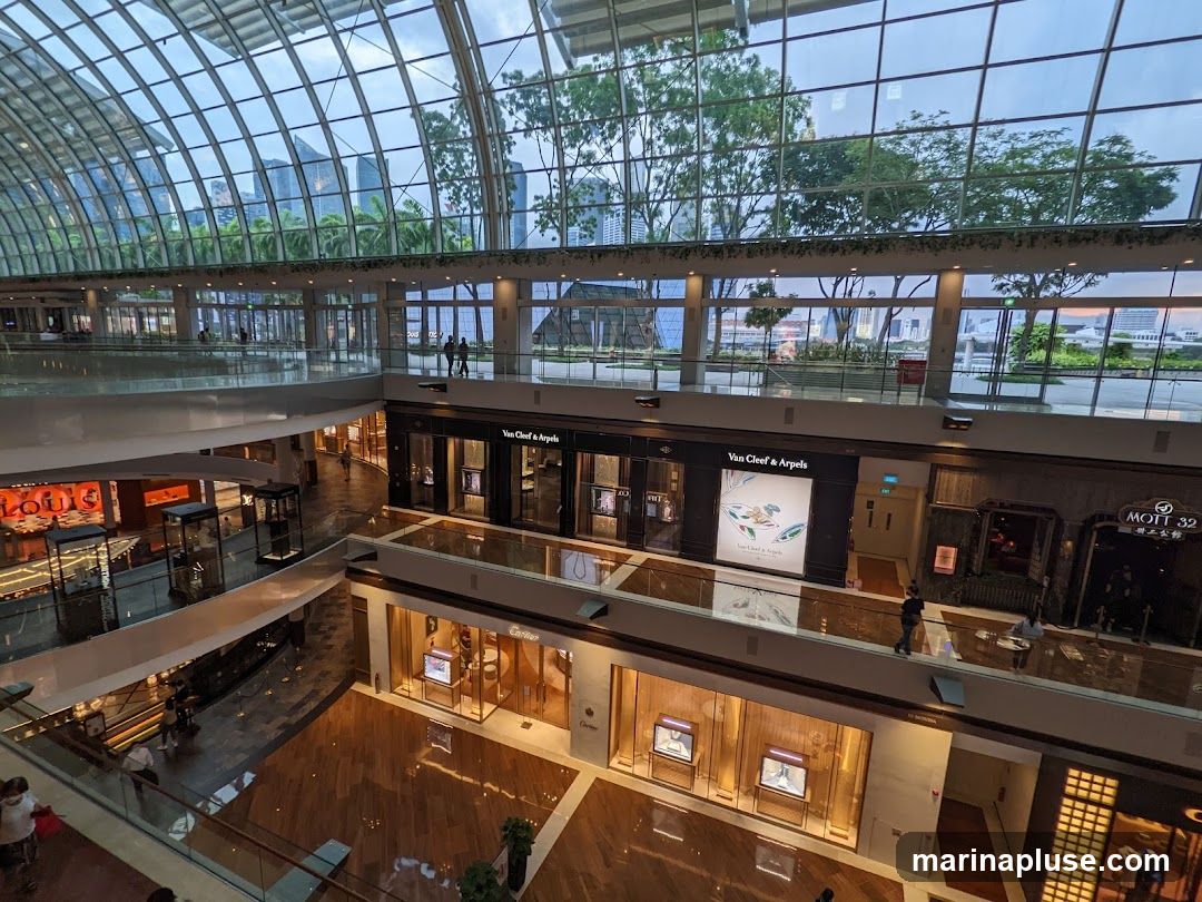 An interior view of a multi-level luxury shopping mall with a glass ceiling and large windows overlooking a cityscape and greenery. Several high-end stores are visible, including Louis Vuitton, Van Cleef & Arpels, Cartier, and Mott 32. The mall features polished wooden floors and glass railings.