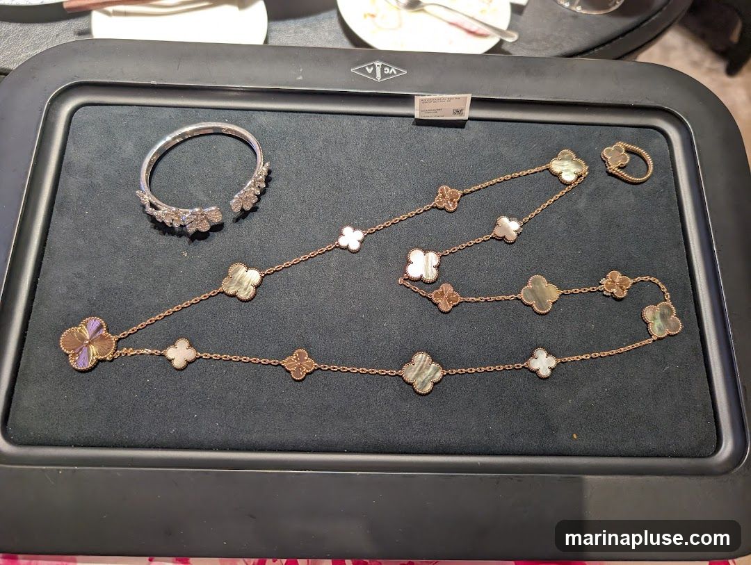 A display of luxury jewelry, including a diamond-encrusted bracelet, a long necklace with clover-shaped motifs in mother-of-pearl and rose gold, and a matching ring. The items are arranged on a dark velvet surface within a display tray.