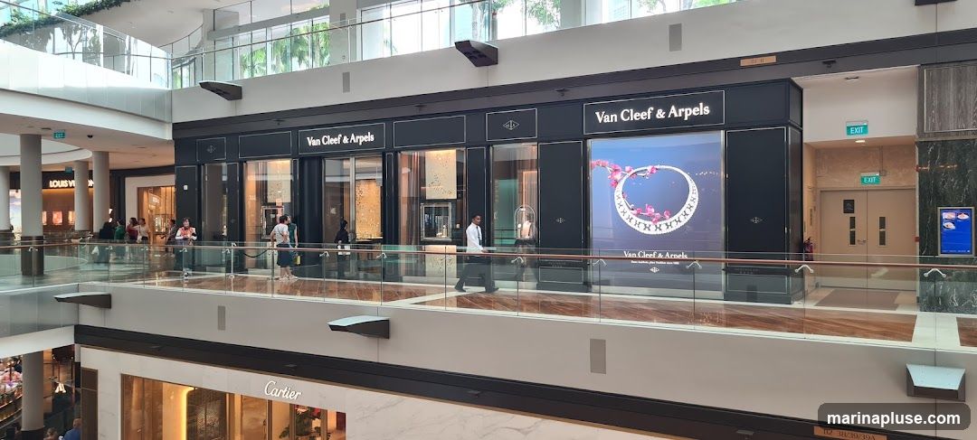 A view of the Van Cleef & Arpels store in a modern shopping mall. The storefront features large display windows showcasing jewelry and a prominent advertisement for a necklace. The brand name "Van Cleef & Arpels" is clearly visible above the windows. Other stores, including Louis Vuitton and Cartier, are visible in the background.