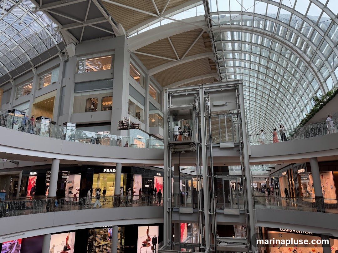 The image shows the interior of a multi-level shopping mall with a distinctive glass and steel roof structure. Several shops are visible on different floors, including Prada and Louis Vuitton, with their storefronts displaying merchandise. An elevator shaft with glass-enclosed elevators is a prominent feature in the center. People are seen browsing and walking on the elevated walkways and balconies. The architecture is sleek and contemporary, with a focus on natural light filtering through the large glass panels of the ceiling.