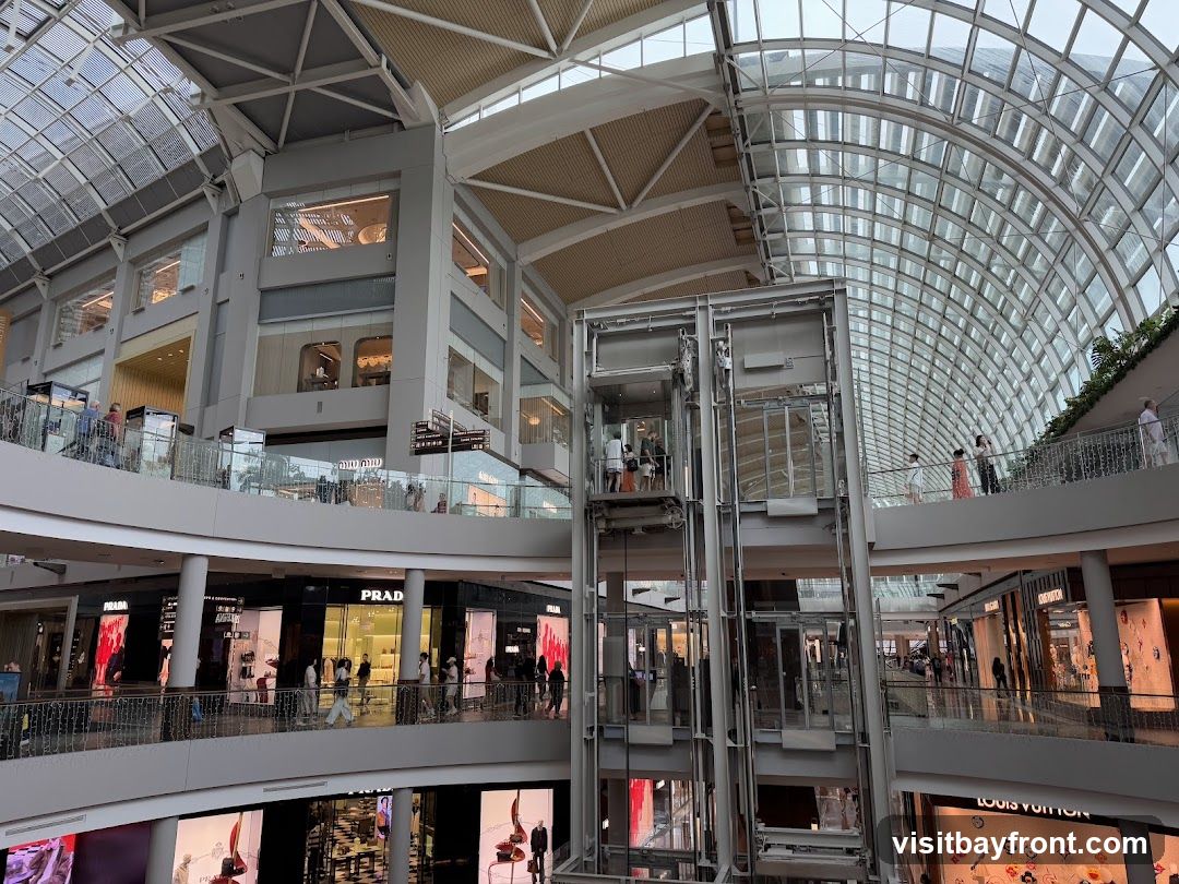 The image shows the interior of a multi-level shopping mall with a distinctive glass and steel roof structure. Several shops are visible on different floors, including Prada and Louis Vuitton, with their storefronts displaying merchandise. An elevator shaft with glass-enclosed elevators is a prominent feature in the center. People are seen browsing and walking on the elevated walkways and balconies. The architecture is sleek and contemporary, with a focus on natural light filtering through the large glass panels of the ceiling.