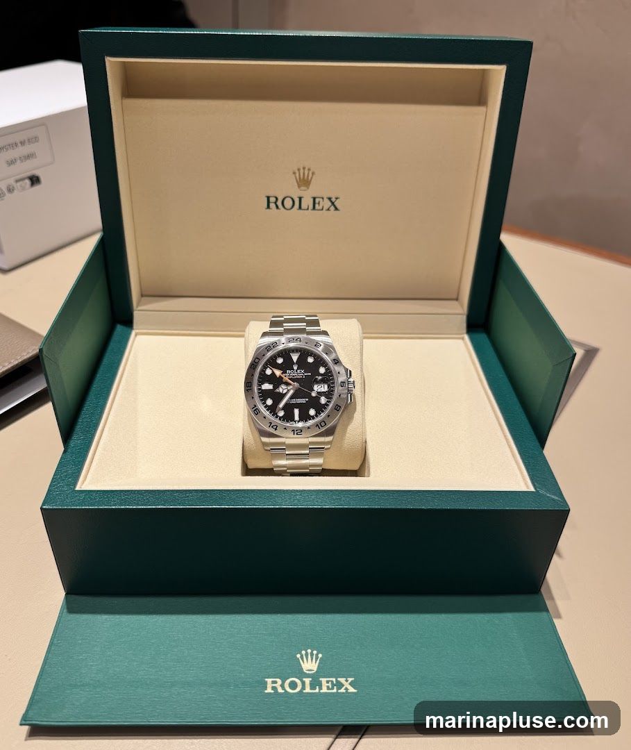A Rolex watch is displayed inside its open green presentation box. The watch has a black dial with silver hour markers and hands, and a stainless steel bracelet. The Rolex logo and name are visible on the inside lid of the box and on the watch face. A white box with text is partially visible in the background.