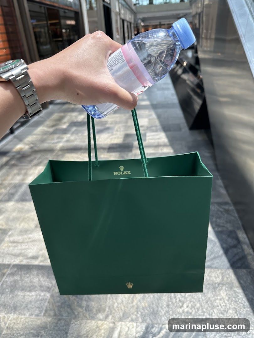 A hand holds a green shopping bag with the Rolex logo and a bottle of Evian water. The bag appears to be made of paper and has green rope handles. The Rolex logo is visible on the front of the bag and on the inside rim. The background shows a tiled floor and what appears to be a shopping mall or arcade.