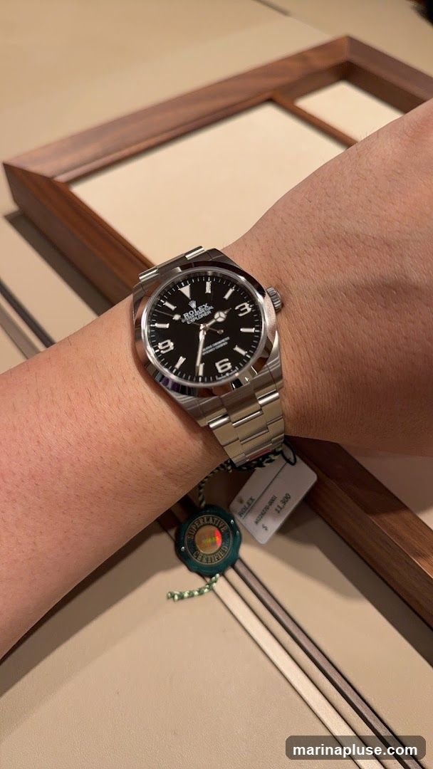 A close-up shot of a Rolex Explorer watch on a person's wrist. The watch has a black dial with white Arabic numerals and markers. A price tag is attached to the watch band, showing a price of $11,300. A green and gold seal with the text "SUPERLATIVE CERTIFIED" is also visible. The background appears to be a display case with a wooden frame.