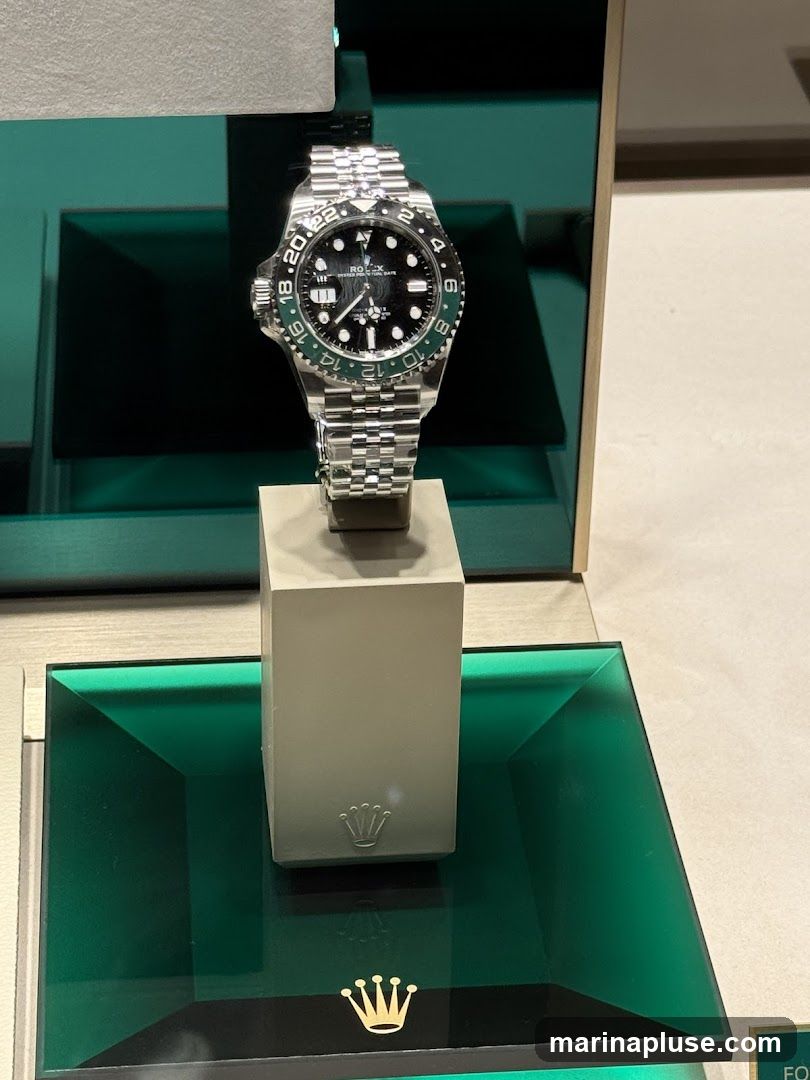 A close-up shot of a Rolex watch with a green bezel and black dial, displayed on a light-colored stand. The watch is made of stainless steel with a jubilee bracelet. The display features green accents and the Rolex crown logo.